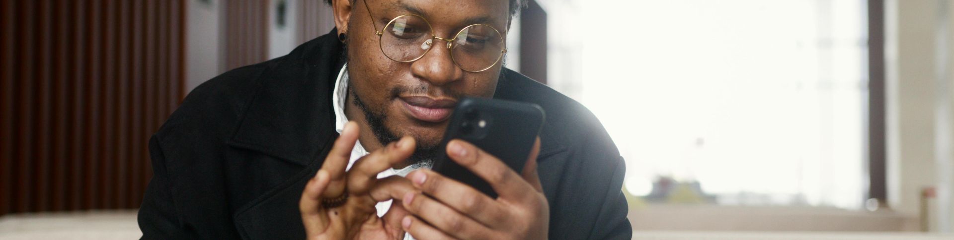 A cheerful man wearing a stylish outfit uses a smartphone while seated in a modern indoor setting.