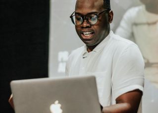 A focused speaker wearing a white shirt, presenting with a laptop on stage during an event.